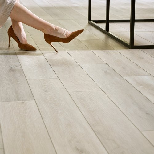 Close-up of luxury vinyl tile (LVT) flooring in a light wood finish, with a person wearing brown high heels seated nearby, showcasing the floor's elegance and durability — ideal for stylish residential spaces.